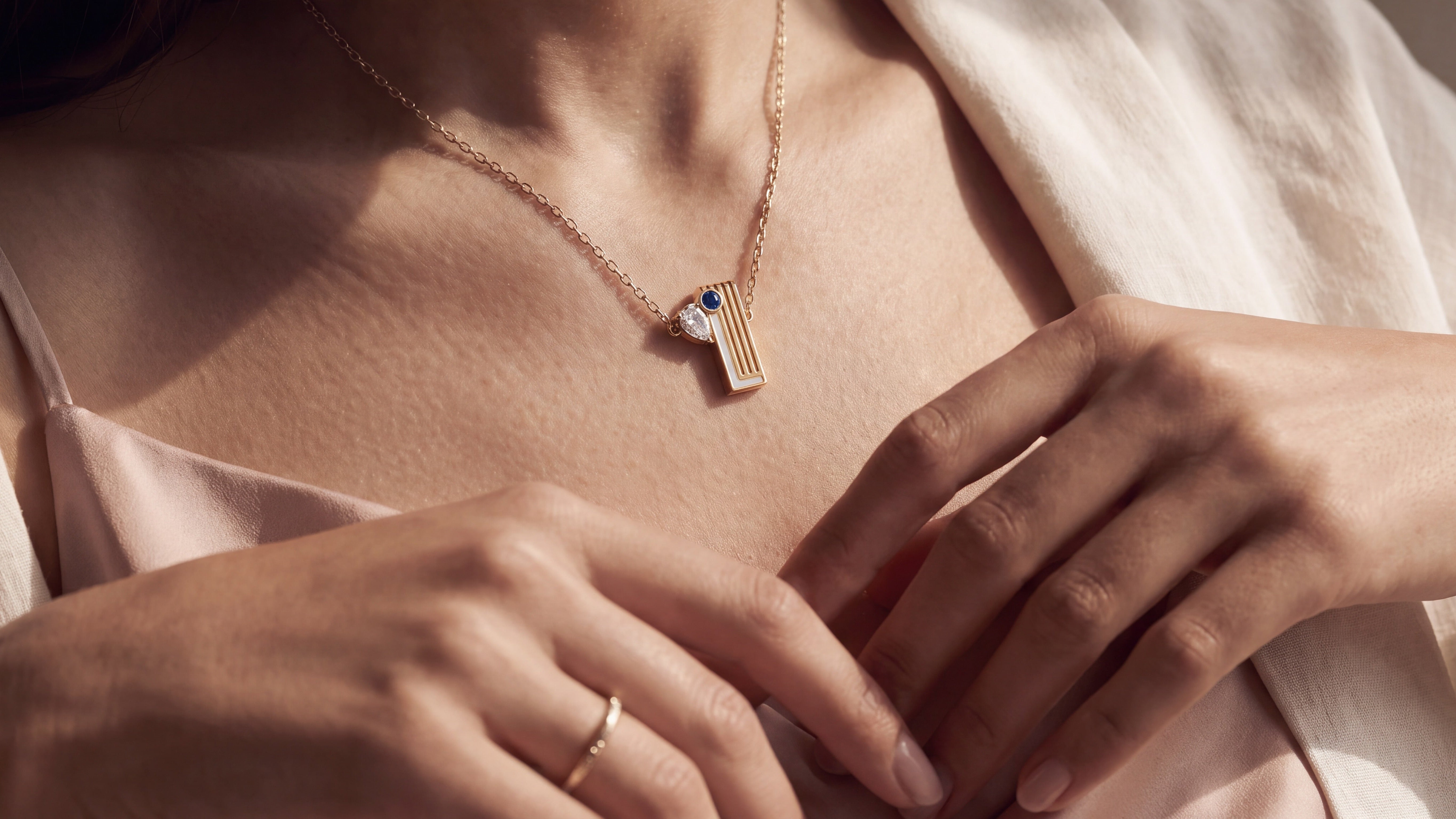 A close-up lifestyle shot of a model wearing a delicate gold necklace with a geometric pendant featuring a blue stone and gold stripes, paired with a soft cream-colored top.