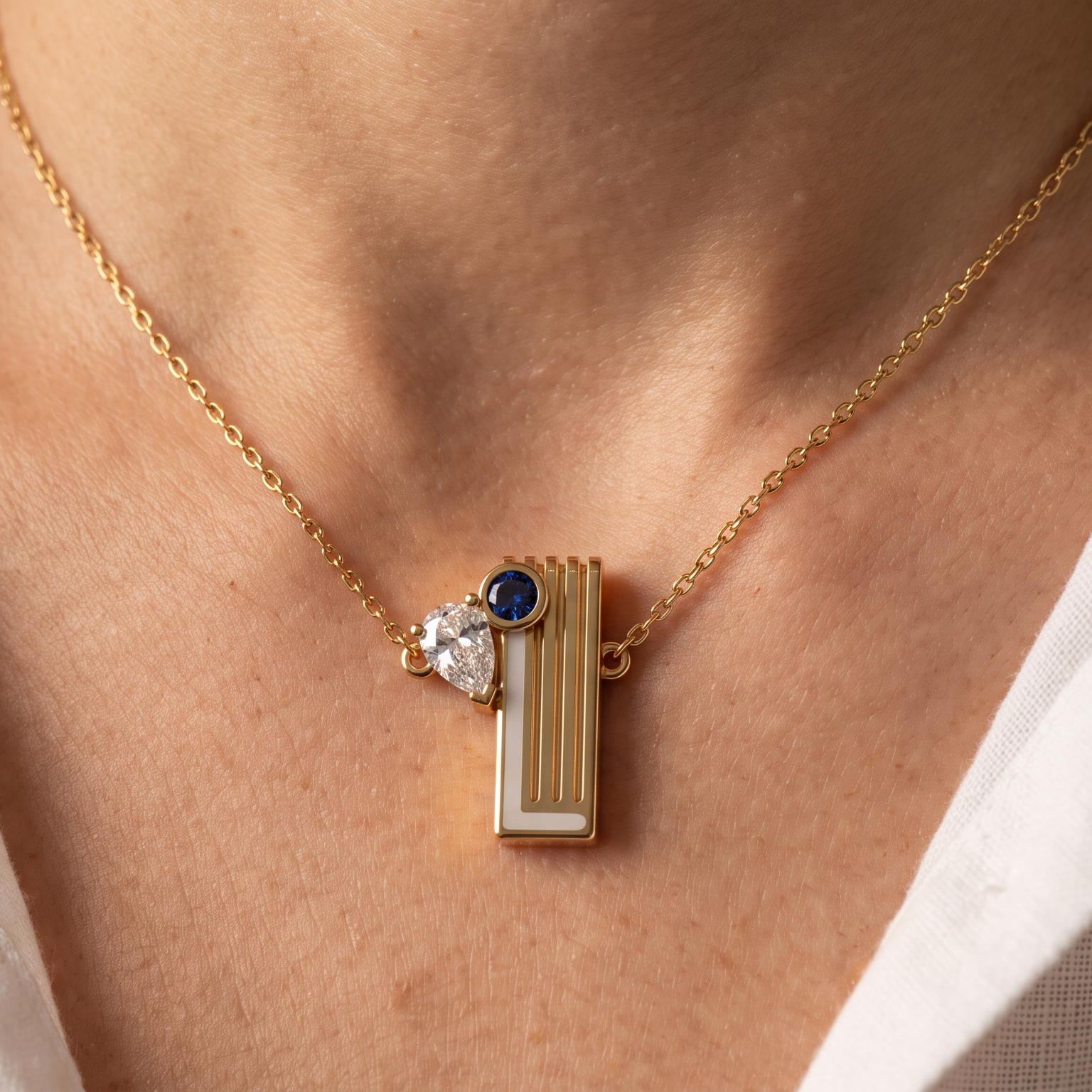 A close-up of a person wearing a delicate gold chain necklace with a geometric pendant featuring a vertical gold bar, a white inlay, a small clear stone, and a dark blue accent stone, styled with a white collared shirt.