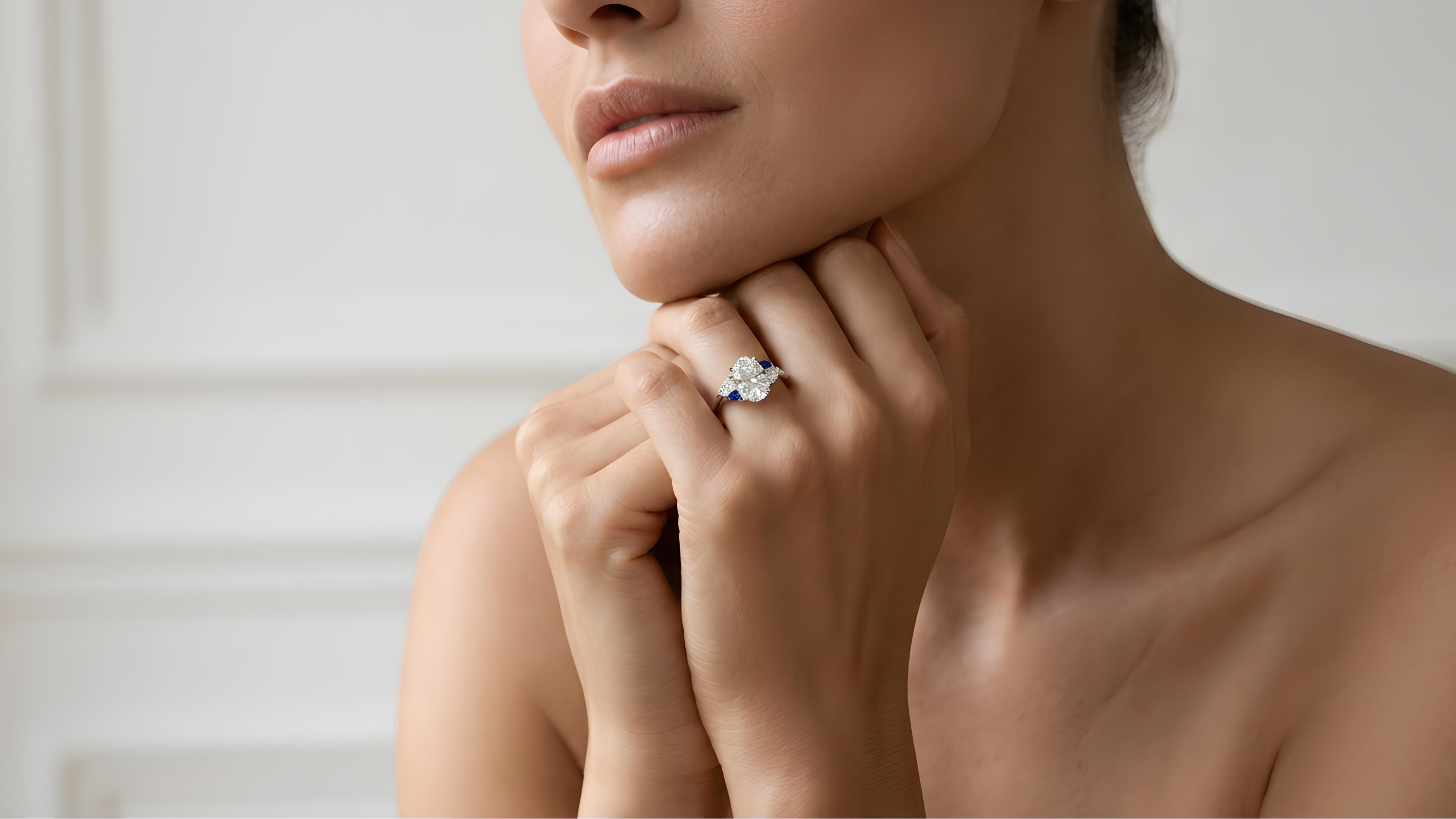 A close-up lifestyle shot of a woman wearing a diamond engagement ring on her finger, with her hand resting on her chin against a soft, light-colored paneled wall.