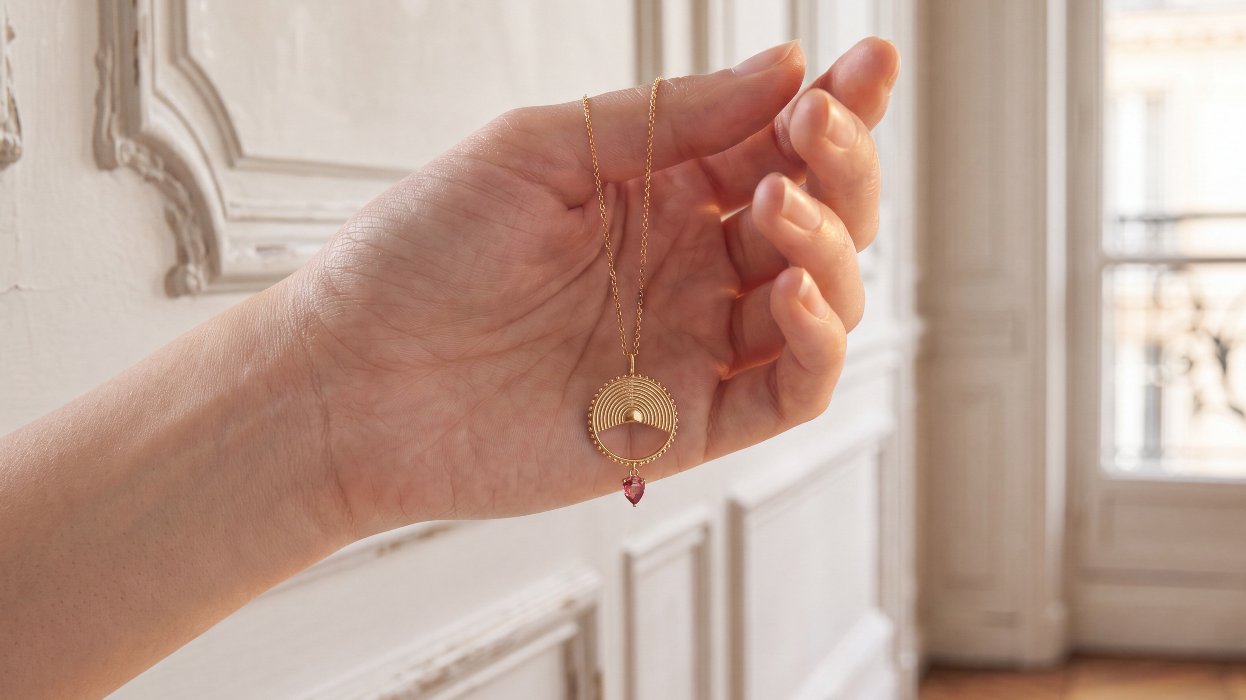 A gold pendant necklace with a crescent moon and central diamond-shaped cutout, held in a person's hand in a bright, elegant room with white paneled walls and a window. with pear shaped pink gemstone