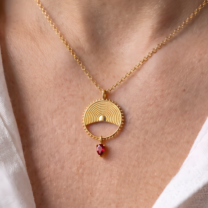 A gold pendant necklace worn by a person, featuring a circular sunburst design with a small red gemstone dangle, set against a white shirt.