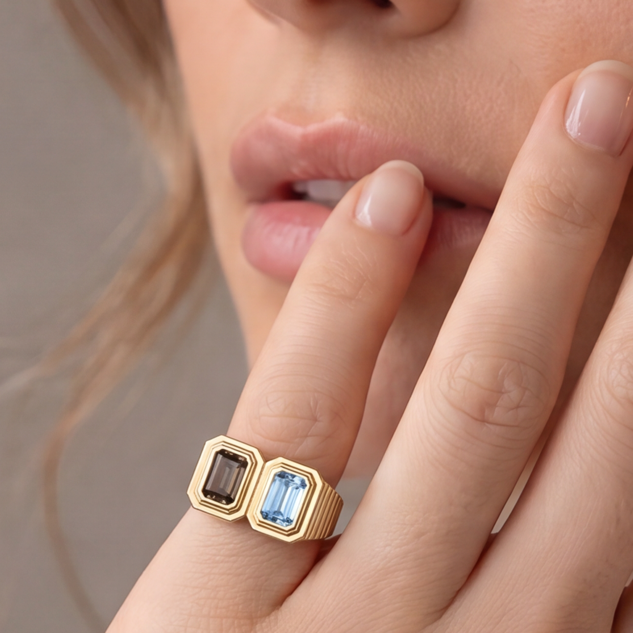 A close-up lifestyle shot of a woman wearing a gold ring with a rectangular light blue gemstone, resting her hand near her face against a neutral gray background.