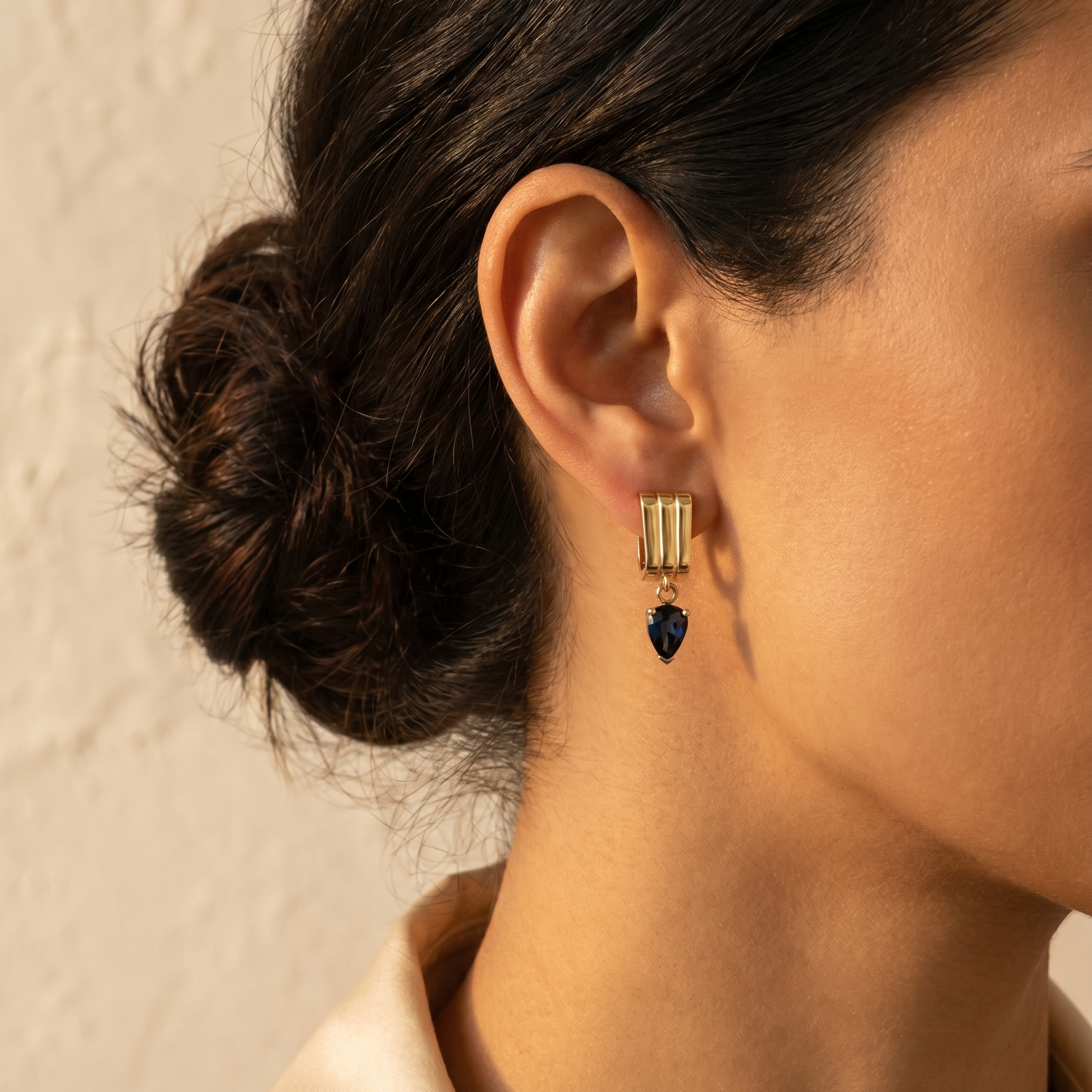 Close-up of a person wearing gold earrings with a dark blue gemstone against a neutral background
